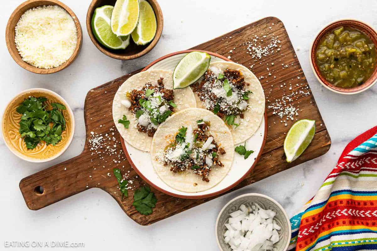 Three street tacos with seasoned ground beef, chopped onions, cilantro, and cheese on corn tortillas are served with lime wedges on a white plate atop a wooden board. Bowls of extra cheese, lime slices, cilantro, onion, and salsa surround the dish.