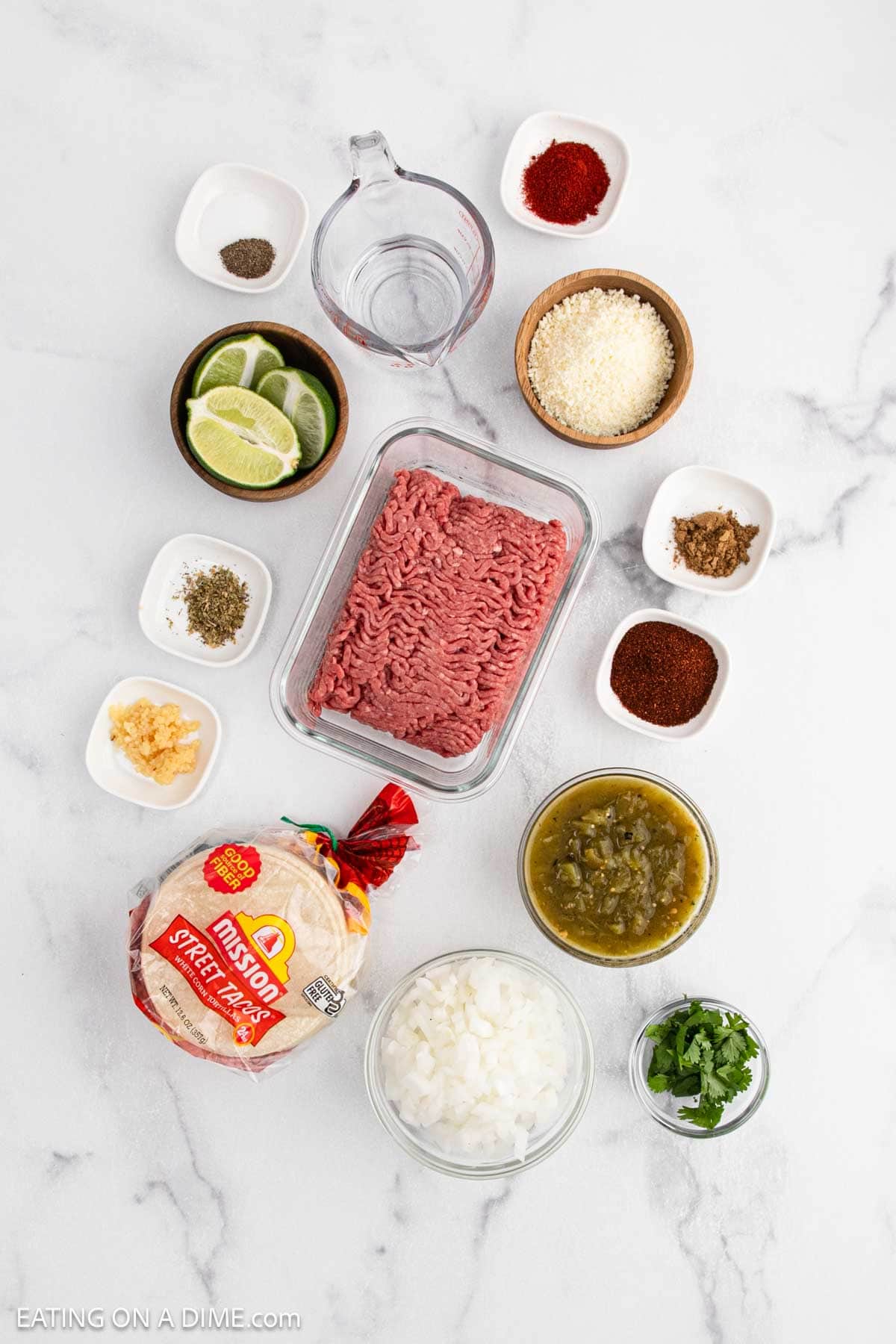 Overhead view of street tacos ingredients on a white surface, including ground beef, tortillas, onion, cilantro, green chilies, lime wedges, seasonings, minced garlic, and grated cheese in small bowls.