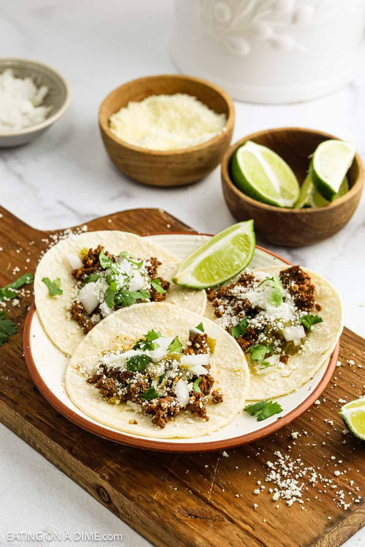 Three street tacos filled with seasoned ground beef, chopped onions, cilantro, and grated cheese are served on a plate with lime wedges. The plate rests on a wooden board, surrounded by bowls of grated cheese, lime slices, and onions.