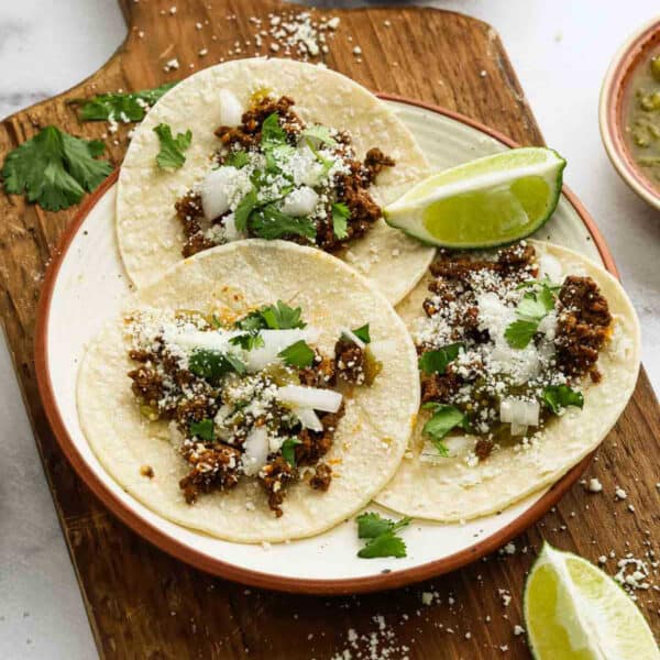 Three street tacos filled with seasoned ground beef, chopped onions, cilantro, and crumbled cheese are served on a plate with lime wedges on a wooden board.