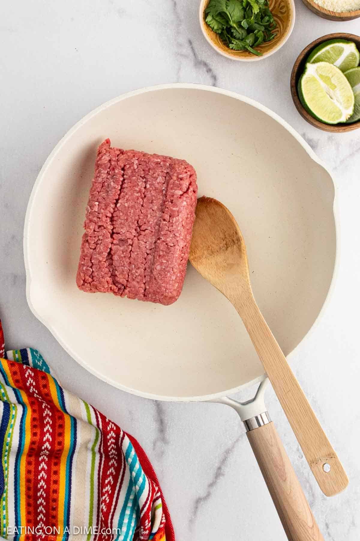 A block of raw ground beef in a white skillet with a wooden spoon, ready to become flavorful beef tacos. Nearby are a colorful striped towel, lime wedges, chopped cilantro, and seasonings for delicious street tacos on a marble surface.