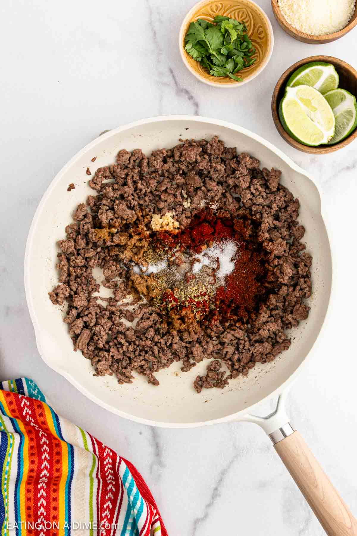 Ground beef for street tacos cooks in a white skillet, topped with spices. Small bowls of chopped cilantro, grated cheese, and lime slices surround the skillet, set against a colorful striped cloth.