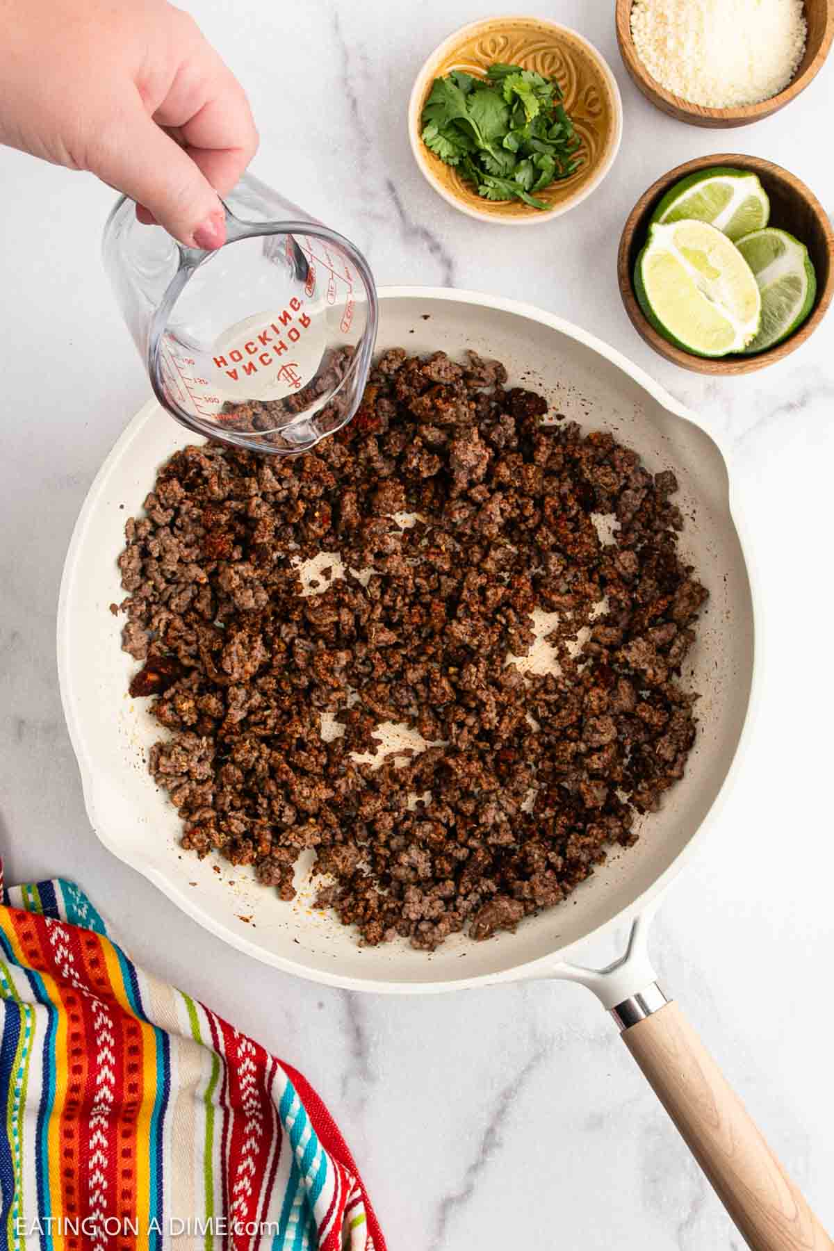 A hand pours water from a measuring cup into a skillet with cooked ground beef for beef tacos. Nearby are bowls of chopped cilantro, grated cheese, lime wedges, and a colorful striped cloth.