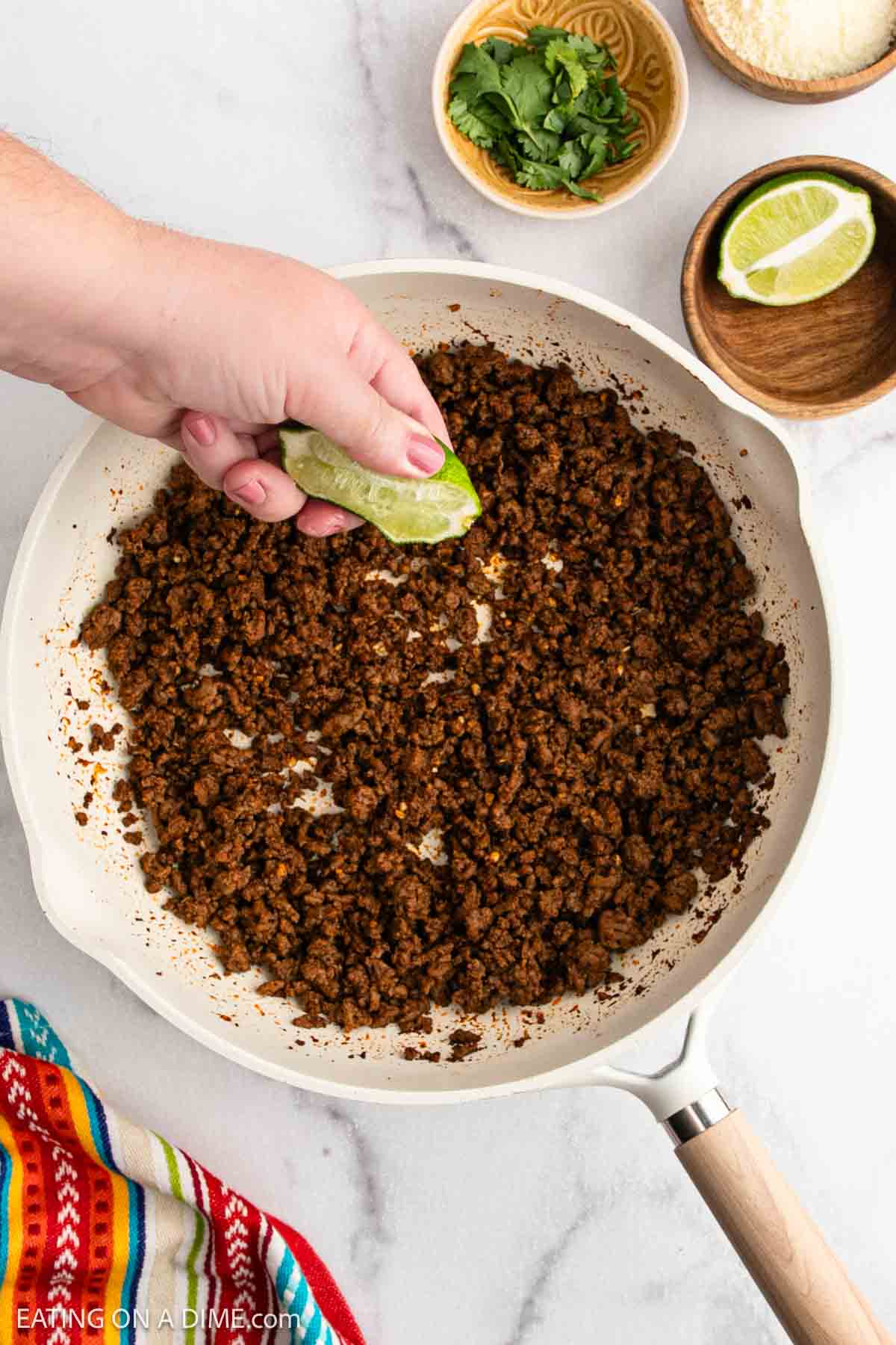 A hand squeezes a lime wedge over a skillet of cooked ground beef, perfect for street tacos. Nearby are small bowls with cilantro, lime, and grated cheese, and a colorful striped cloth is partially visible.