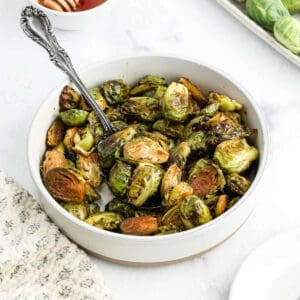 A white bowl filled with Honey Balsamic Brussels Sprouts, some halved and browned, with a silver spoon resting inside. The bowl sits on a white surface next to a patterned cloth.