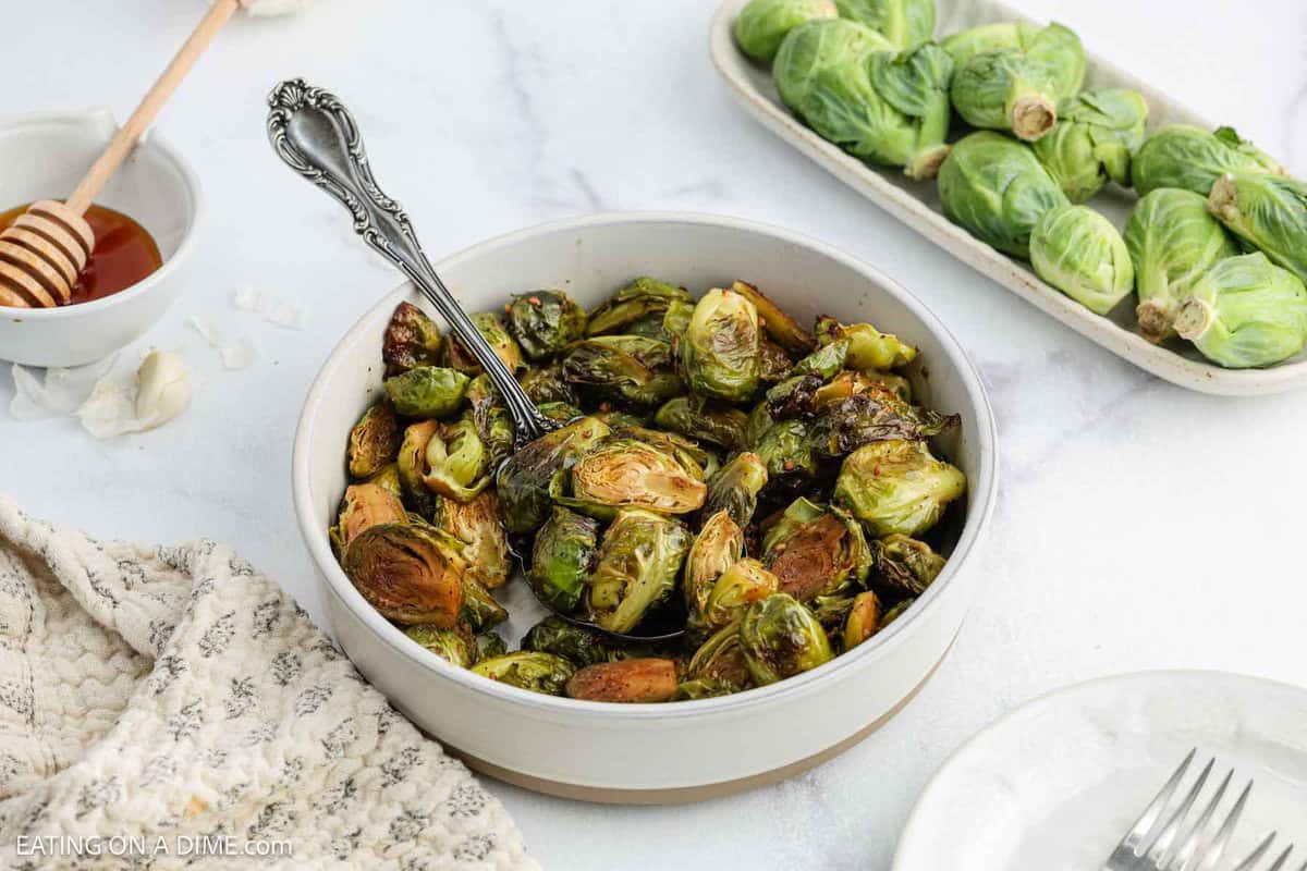 A round dish filled with Honey Balsamic Brussel Sprouts with a serving spoon, next to a plate of fresh Brussels sprouts, a small bowl of honey with a honey dipper, and a fork and napkin on a white surface.