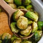 A wooden spoon holds several roasted Honey Sriracha Brussels sprouts, showing their browned, crispy edges. More Brussels sprouts are visible in a bowl in the background.