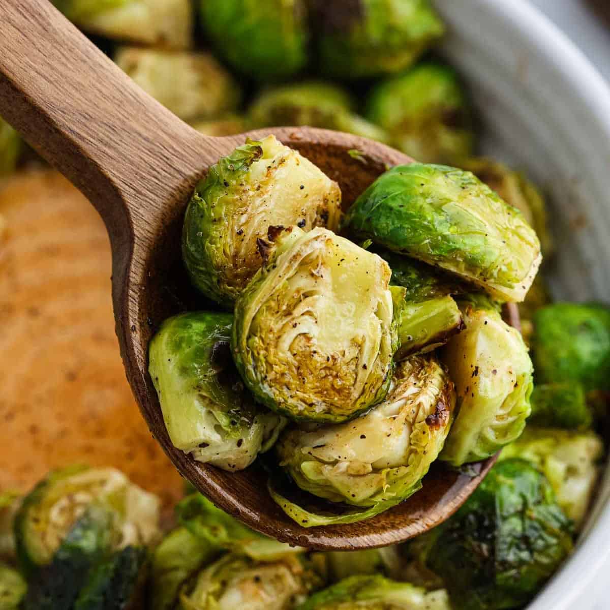 A wooden spoon holds several roasted Honey Sriracha Brussels sprouts, showing their browned, crispy edges. More Brussels sprouts are visible in a bowl in the background.