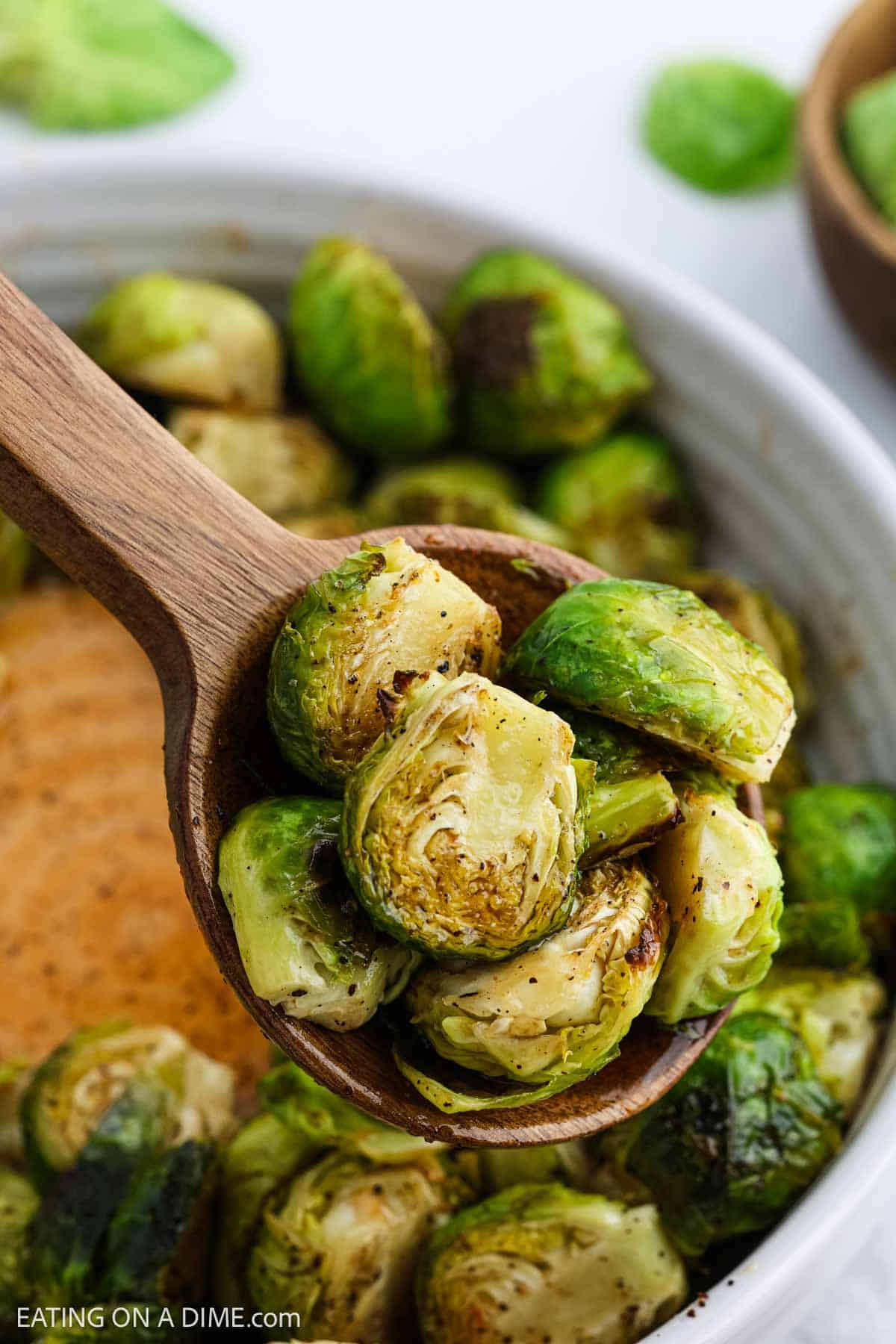 A wooden spoon holds Honey Sriracha Brussel Sprouts above a bowl filled with more golden brown, lightly seasoned vegetablesโan irresistible twist on a classic Brussel Sprouts recipe.