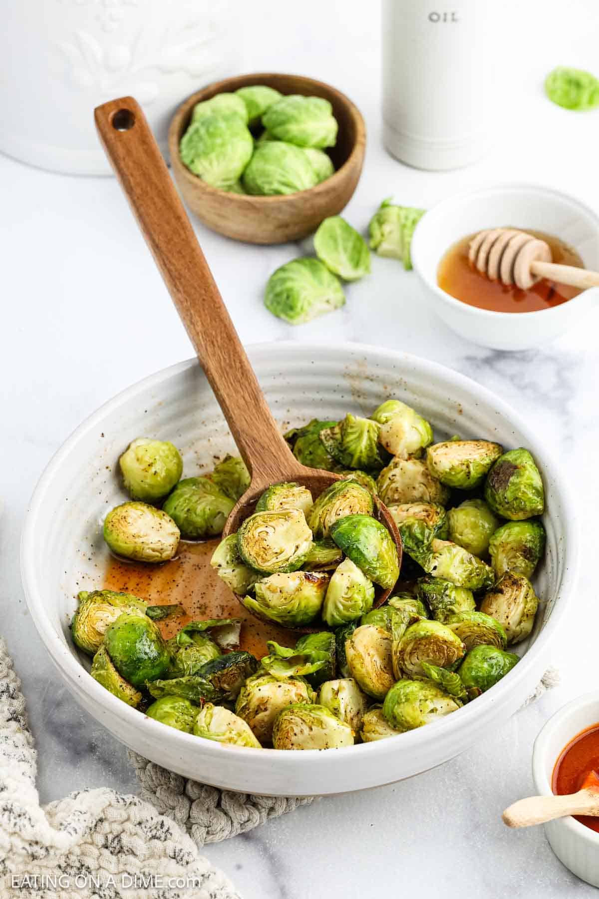 A white bowl filled with Honey Sriracha Brussels sprouts, some tossed in a wooden spoon. Fresh Brussels sprouts, a small dish of honey with a dipper, and a bottle labeled "oil" surround the bowl for a burst of sweet heat.