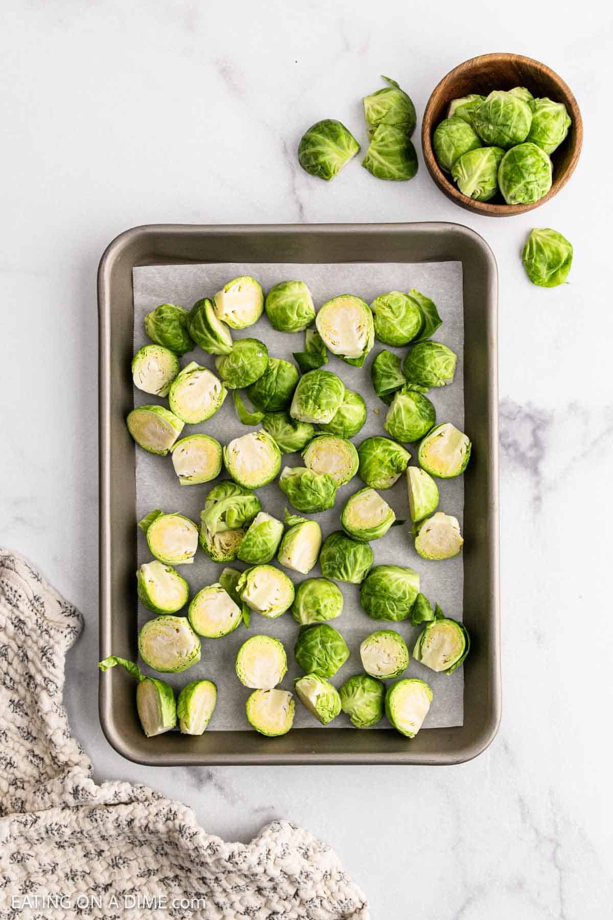 A baking sheet lined with parchment paper holds halved raw Brussels sprouts, ready to become Honey Sriracha Brussel Sprouts. A small bowl of extra Brussels sprouts sits in the upper right corner, with a textured cloth on the marble surface below.