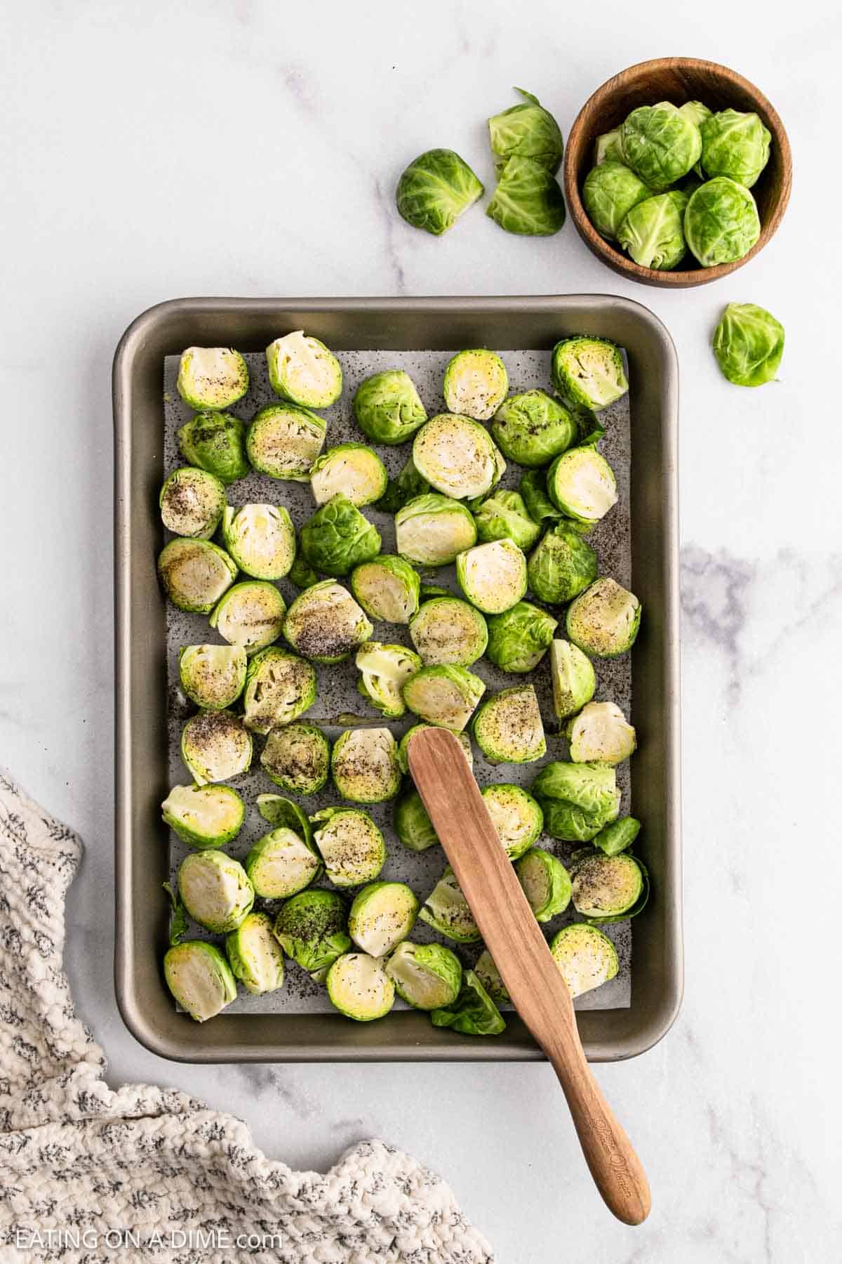 A baking sheet with halved raw Brussels sprouts seasoned for Honey Sriracha Brussel Sprouts, a wooden spatula, a small bowl of extra sprouts, and a textured cloth on a white marble surface.
