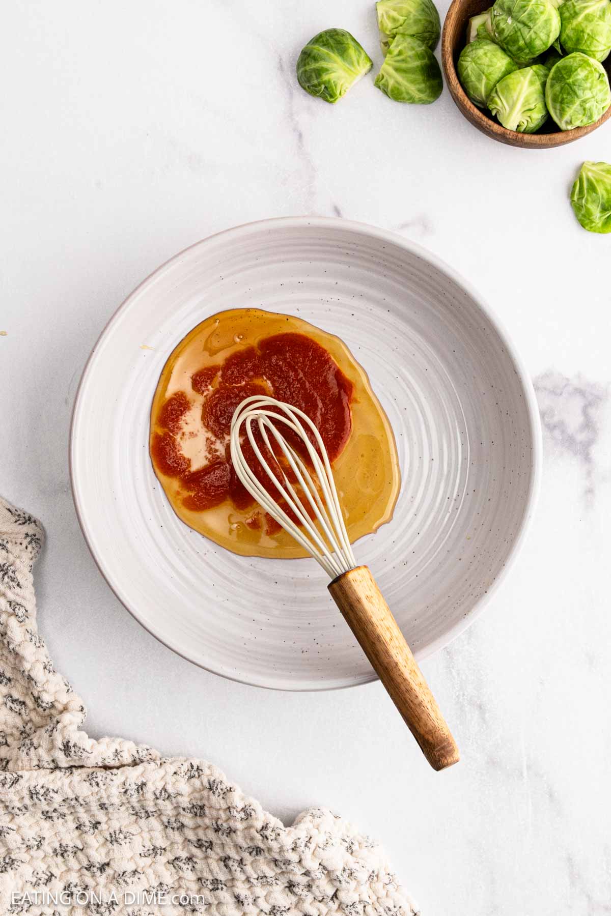 A white bowl with a metal whisk mixes a reddish-brown Honey Sriracha sauce, placed on a marble surface. A small bowl and loose Brussel Sprouts are nearby, with part of a textured gray cloth visible in the corner.