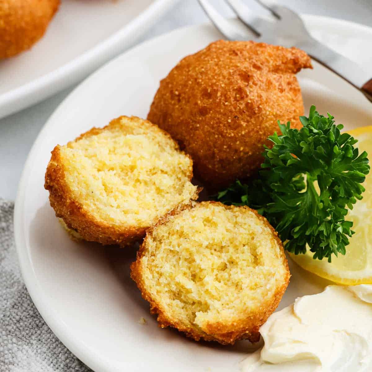 A plate with golden-brown hush puppies, one cut in half to show the moist inside, garnished with parsley and a lemon wedge, perfectly highlighting this classic Southern recipe and served with a dollop of creamy sauce.