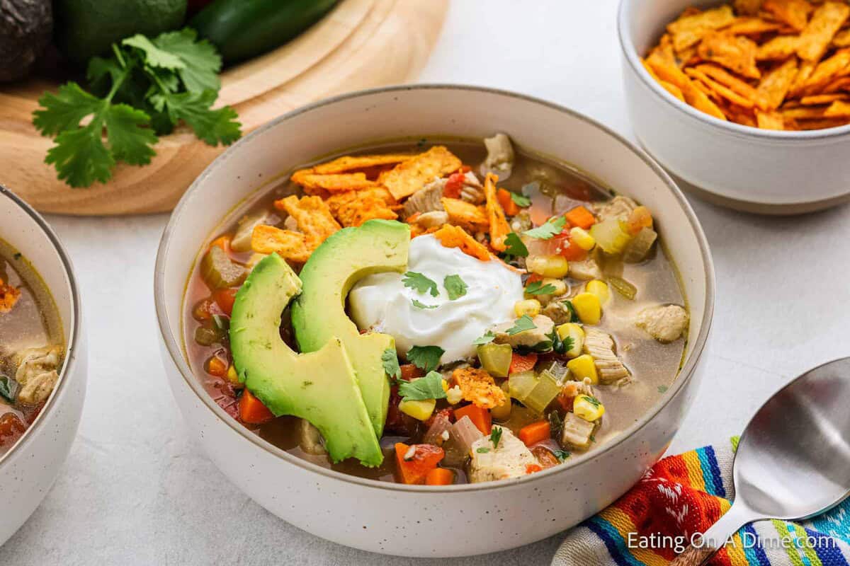 A bowl of Mexican Chicken Soup topped with tortilla chips, avocado slices, sour cream, and cilantro. This vibrant soup is packed with vegetables and served alongside a spoon and a colorful napkin for a festive touch.