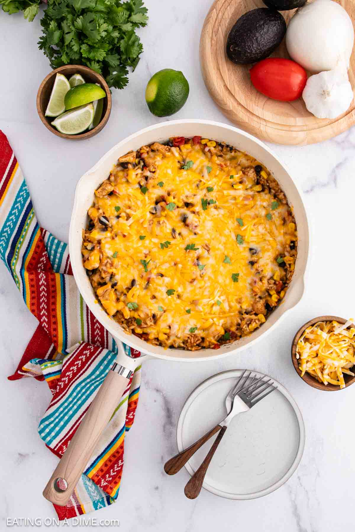 A round casserole dish filled with cheesy baked Mexican Rice Casserole, surrounded by a colorful striped napkin, a plate with forks, a small bowl of shredded cheese, and fresh ingredients like lime, cilantro, avocado, tomato, and garlic.
