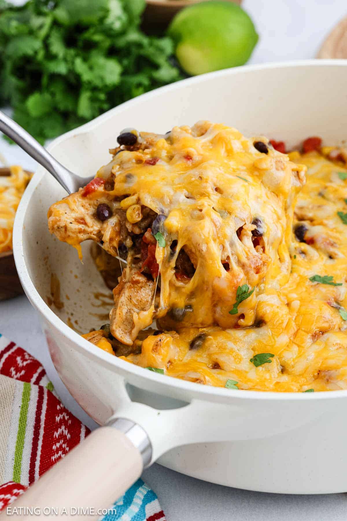A white skillet filled with a cheesy Mexican Chicken casserole mixed with black beans, corn, and diced tomatoes. A serving spoon lifts a portion, showing melted cheese stretching. Fresh cilantro and a lime are in the background.