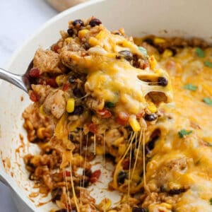 A spoon lifts a cheesy, baked Mexican Chicken and Rice Casserole from the dish, revealing melted cheese, chicken, black beans, corn, rice, and diced tomatoes.