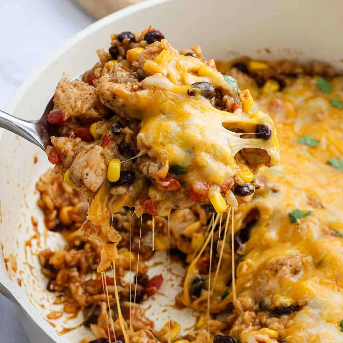 A spoon lifts a cheesy, baked Mexican Chicken and Rice Casserole from the dish, revealing melted cheese, chicken, black beans, corn, rice, and diced tomatoes.