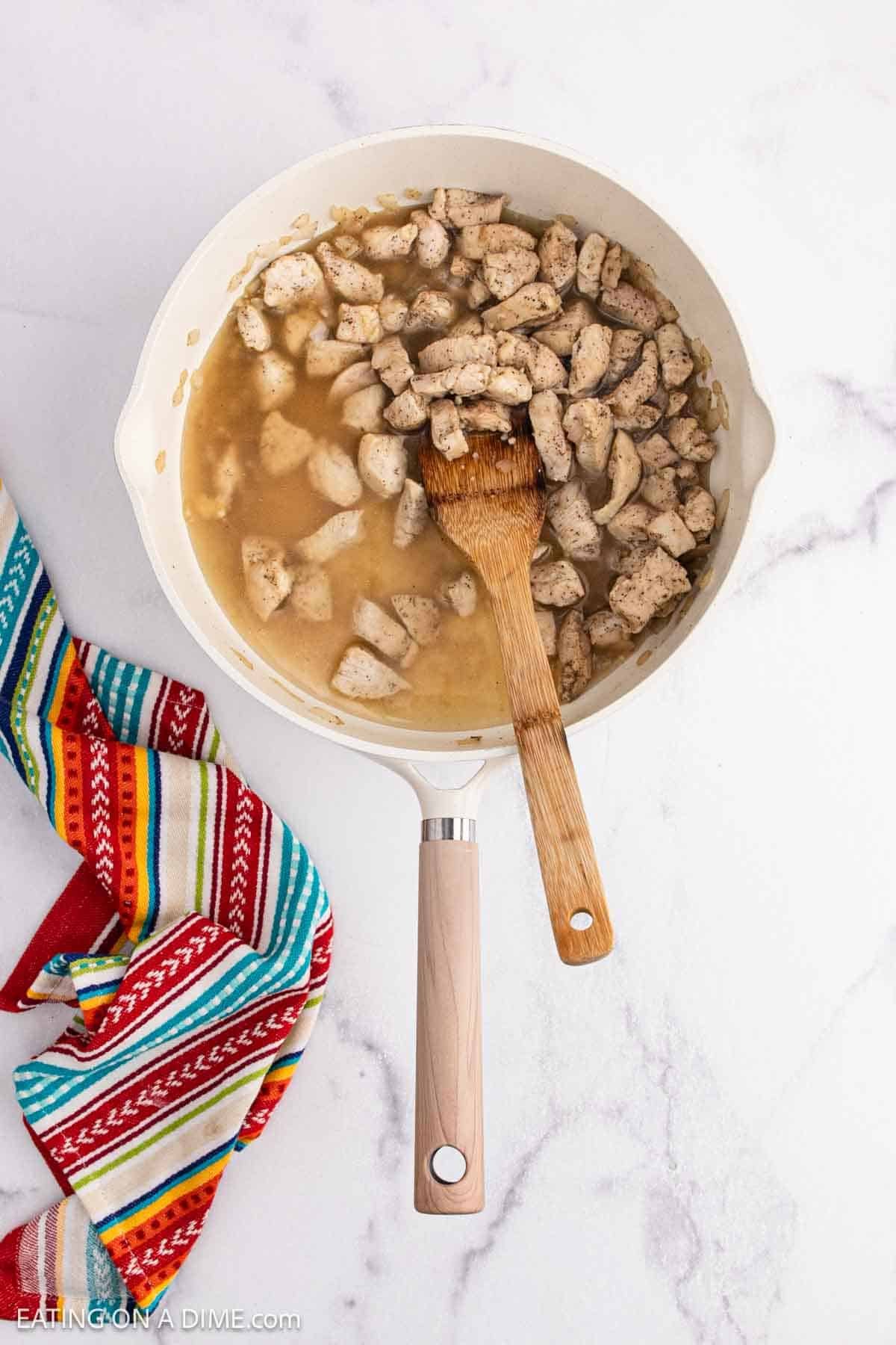 A white skillet filled with cooked, seasoned Mexican Chicken pieces in a light sauce is being stirred with a wooden spoon. A colorful striped towel sits beside the skillet on a white marble surface.