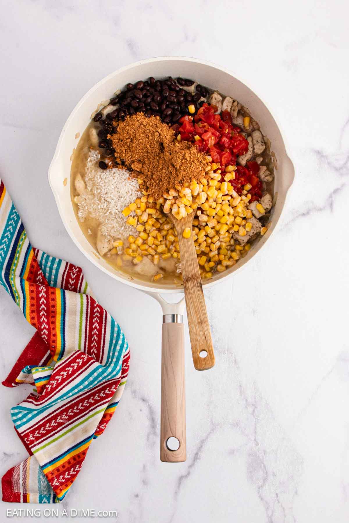A skillet on a white marble countertop holds a flavorful Mexican Chicken and Rice casserole with black beans, corn, diced tomatoes, taco seasoning, and chicken. A wooden-handled spoon rests inside, with a colorful striped cloth beside the pan.