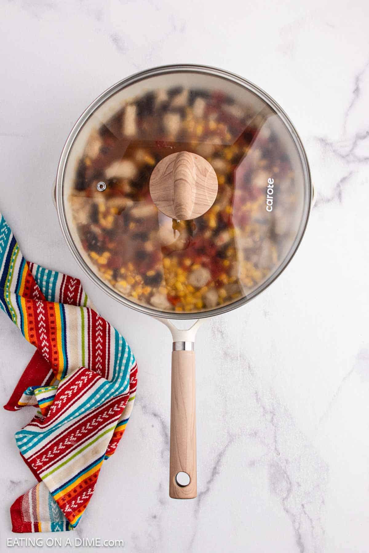 A skillet with a glass lid sits on a white marble surface next to a colorful striped cloth. Inside, the visible mixture of food, including corn and other ingredients, hints at a flavorful Mexican Casserole ready to be enjoyed.