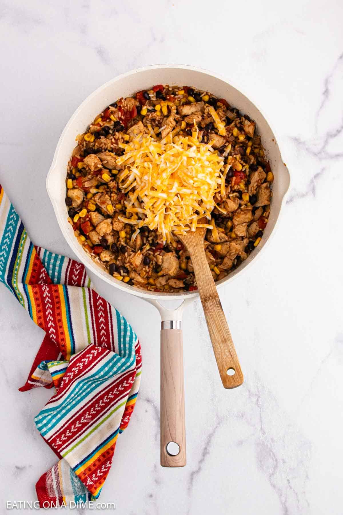 A skillet filled with a colorful Mexican Chicken and Rice casserole—tender chicken, black beans, corn, diced tomatoes, and shredded cheese. A wooden spoon rests inside the pan, with a striped cloth beside it on a white marble surface.