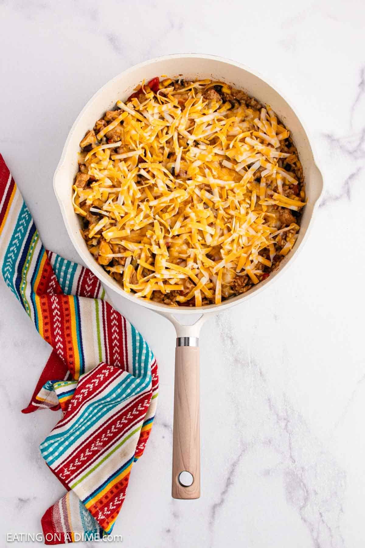 A skillet filled with a cheesy rice casserole topped with shredded cheese sits on a white marble surface, next to a colorful striped kitchen towel.