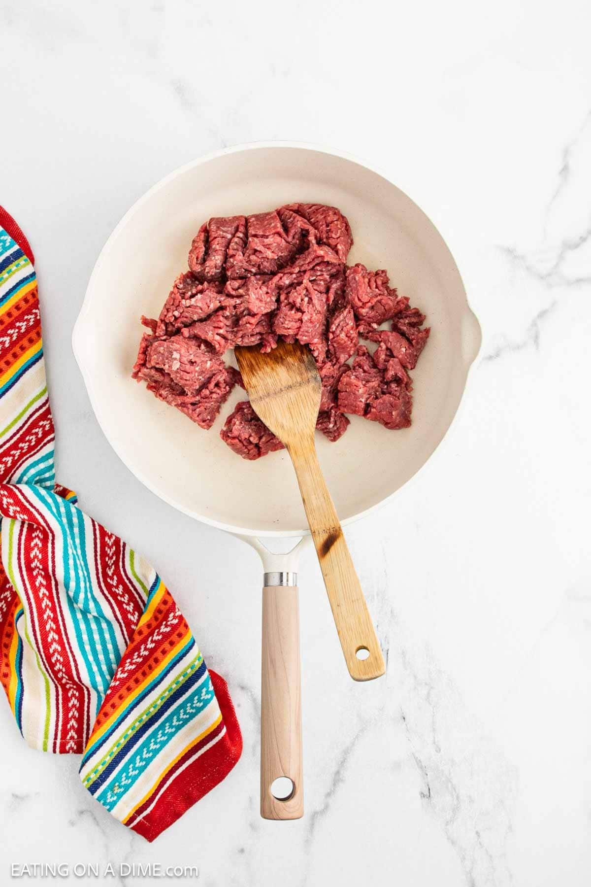 Raw ground beef in a white skillet with a wooden spoon resting on it, ready to be used in a flavorful Mexican Cornbread Casserole. A colorful striped kitchen towel sits to the left on a white marble surface.
