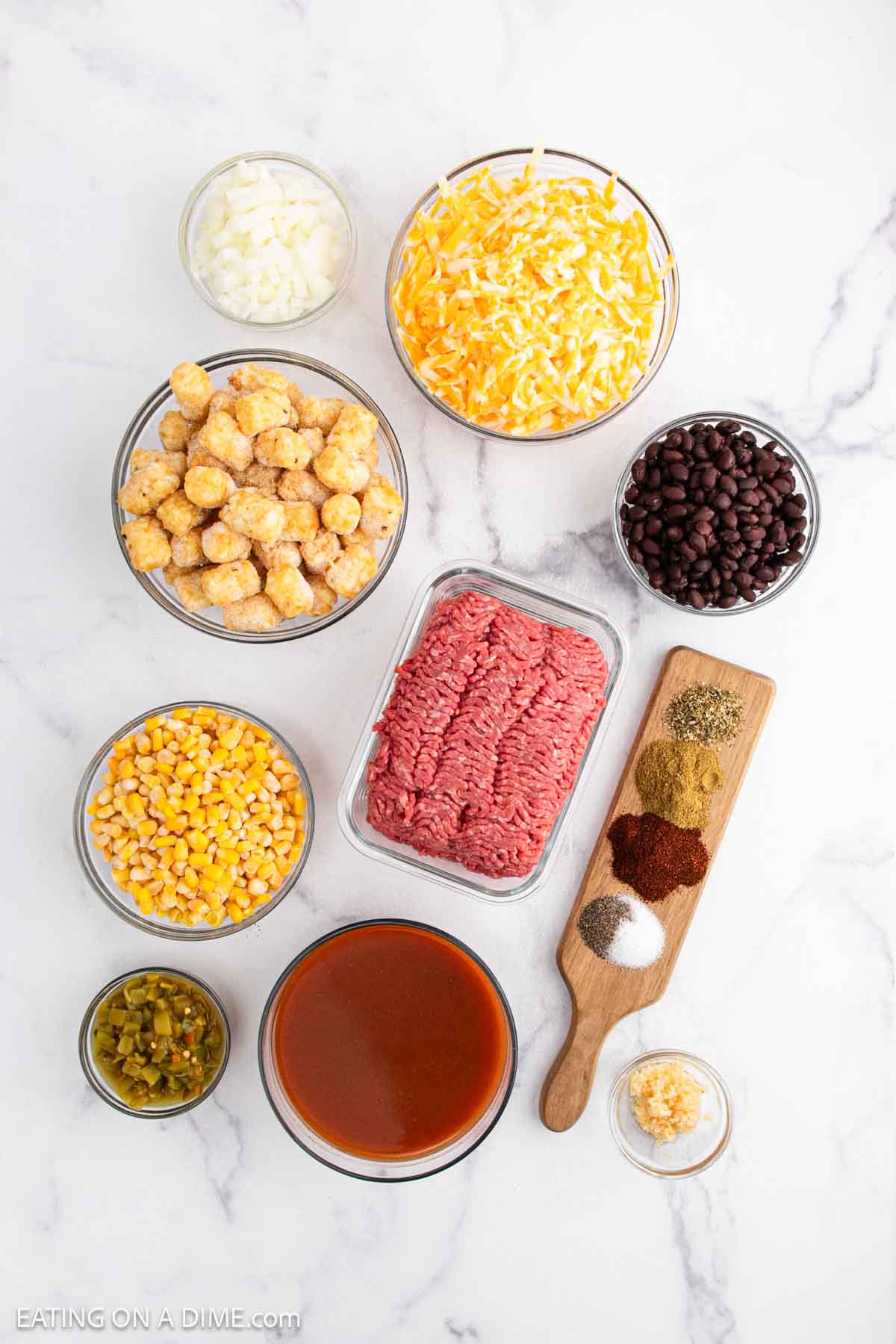 Assorted ingredients for Mexican Tator Tot Casserole on a white marble surface, including bowls of shredded cheese, tater tots, black beans, corn, green chiles, enchilada sauce, ground beef, minced garlic, and a variety of spices.