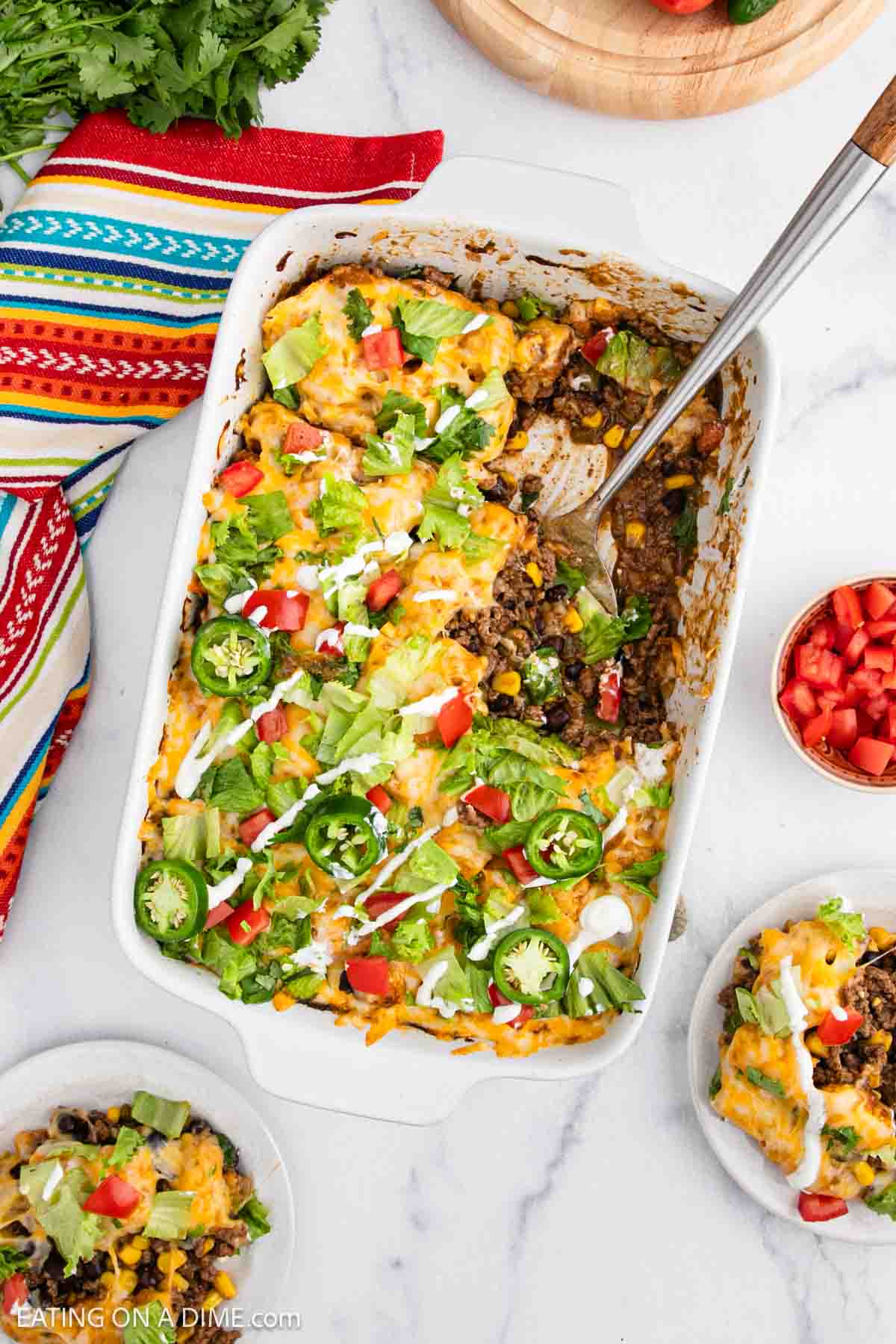 A casserole dish filled with a Mexican Tator Tot Casserole, topped with melted cheese, diced tomatoes, jalapeños, avocado, and fresh greens. Serving spoon inside, two plates nearby, plus a colorful napkin and chopped veggies on the side.