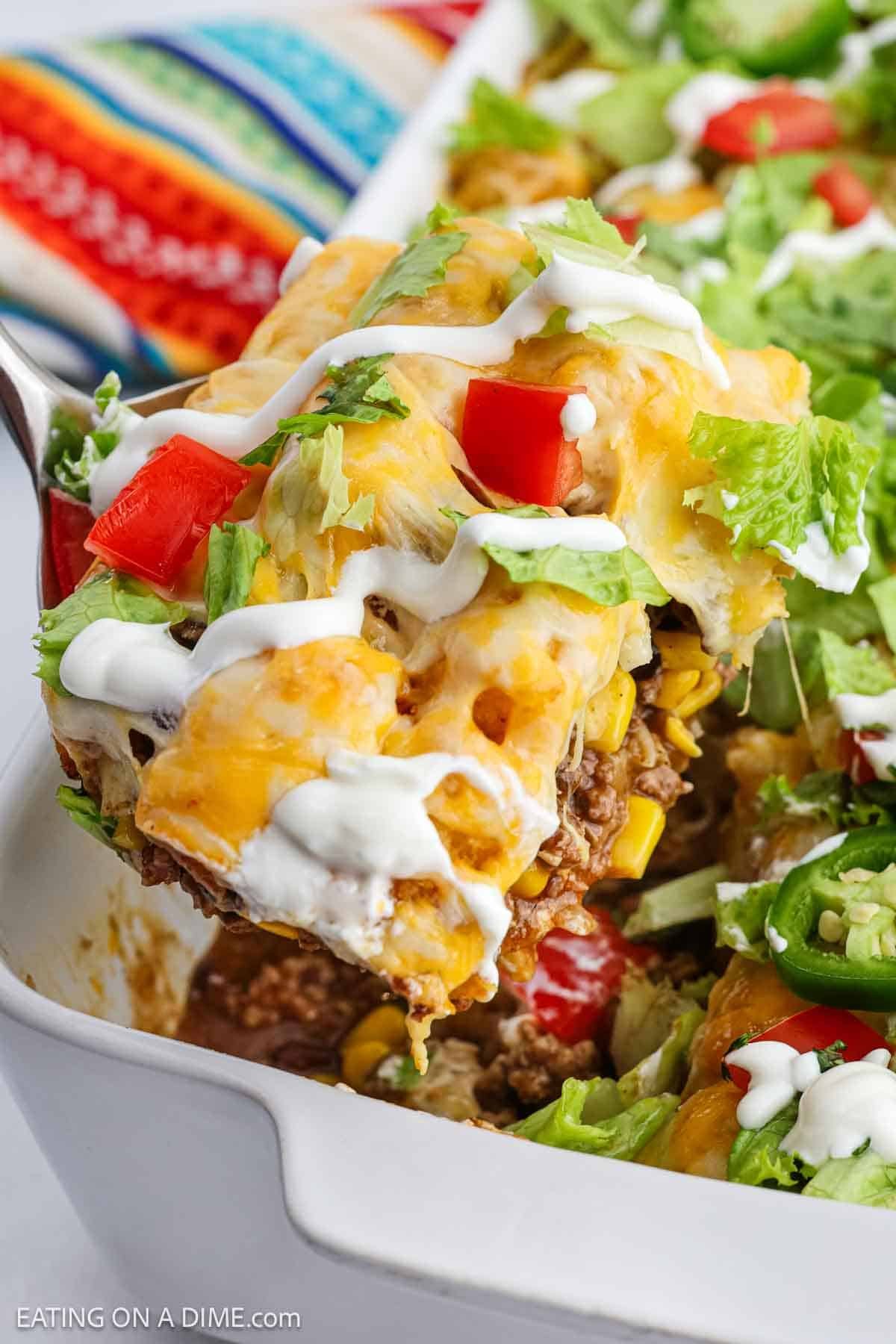 A close-up of a spatula lifting a cheesy Mexican Tator Tot Casserole topped with lettuce, tomatoes, jalapeños, and sour cream from a white baking dish. This colorful Mexican recipe sits on a striped cloth.