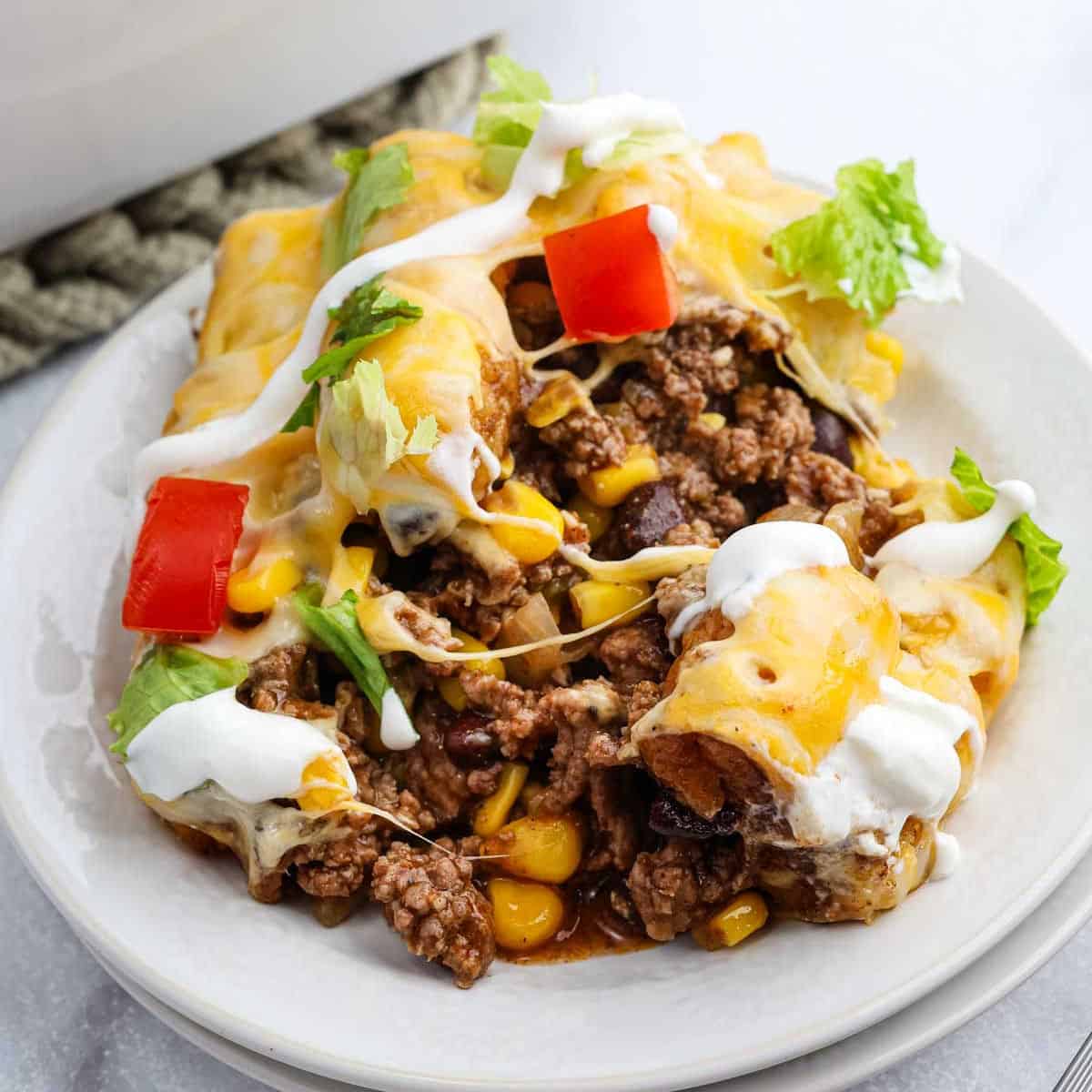 A plate of beef and cheese enchiladas, inspired by a classic Mexican recipe, topped with sour cream, lettuce, tomato, and melted cheese, with ground beef, corn, and beans spilling out of the tortillas.