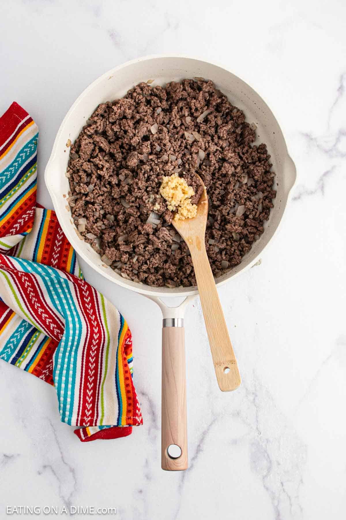 A skillet filled with cooked ground beef and onions sits on a marble countertop, ready for a tasty Mexican Tator Tot Casserole. A wooden spoon holds minced garlic, and a colorful striped kitchen towel rests beside the pan.
