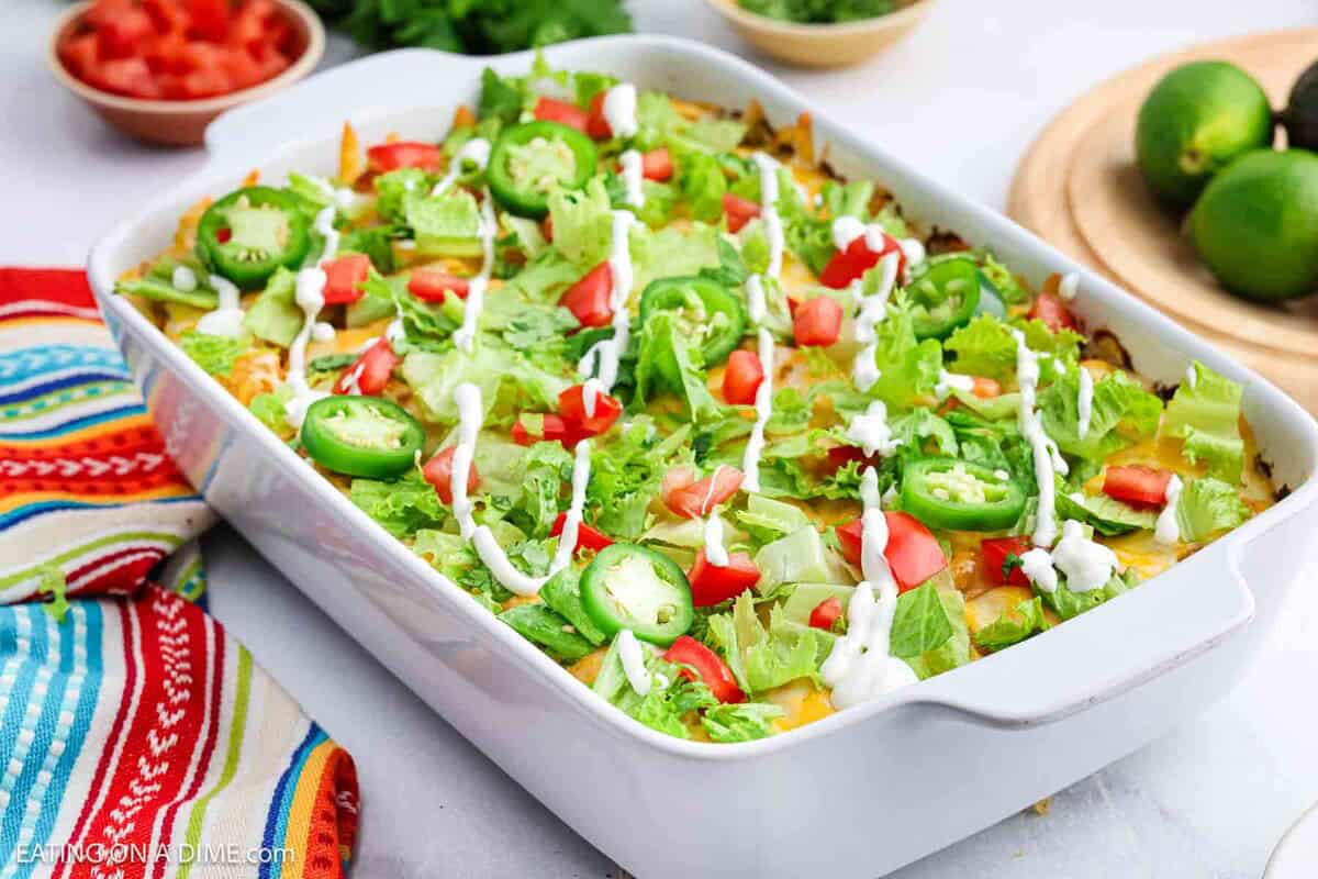 A casserole dish filled with a layered Mexican Tator Tot Casserole topped with lettuce, diced tomatoes, sliced jalapeños, and drizzled with sour cream. Fresh limes, chopped tomatoes, and a colorful striped towel are nearby.