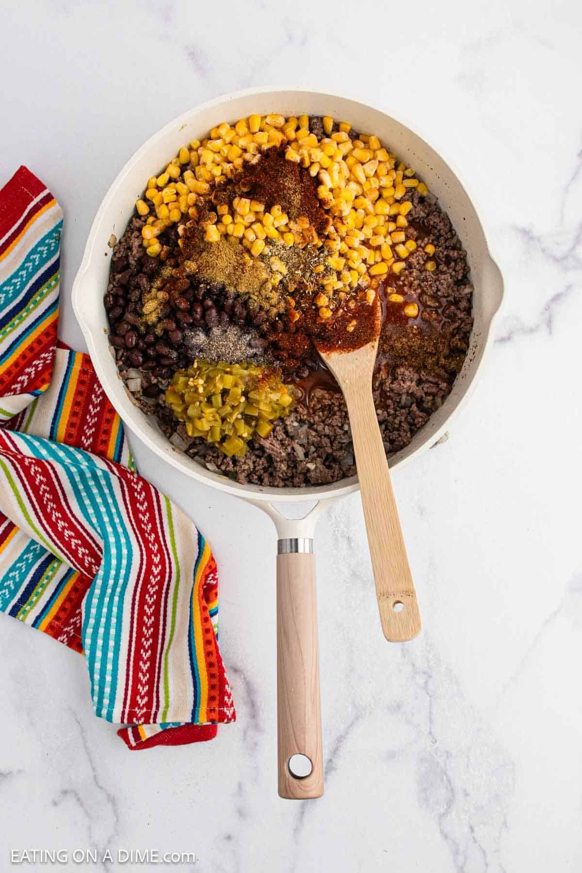 A skillet filled with cooked ground beef, black beans, corn, diced green chilies, and seasonings, perfect for a Mexican casserole, sits on a marble surface next to a colorful striped towel. A wooden spoon is resting in the mixture.