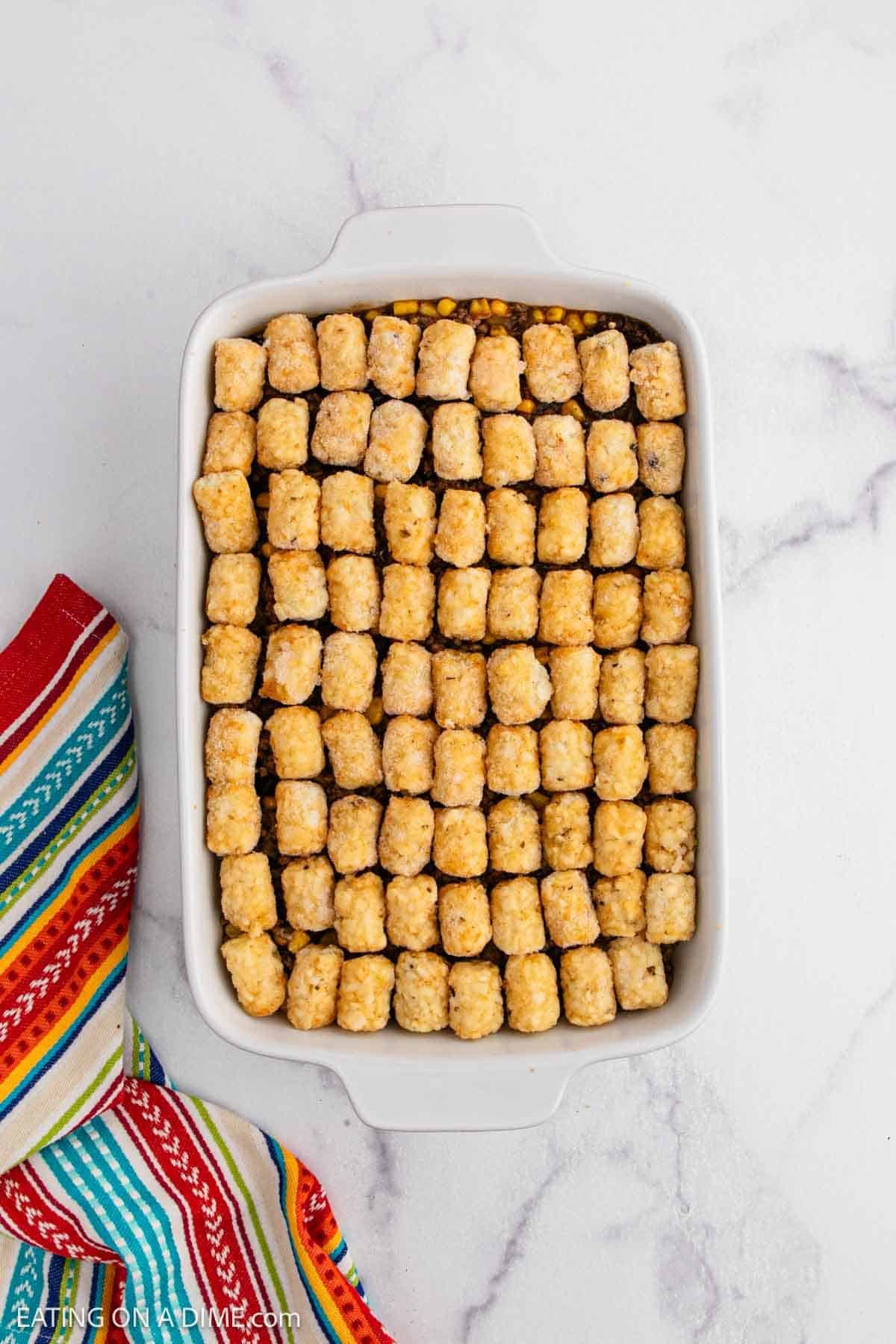 A white baking dish filled with neatly arranged tater tots atop a Mexican Tator Tot Casserole sits next to a colorful striped cloth on a white marble surface.