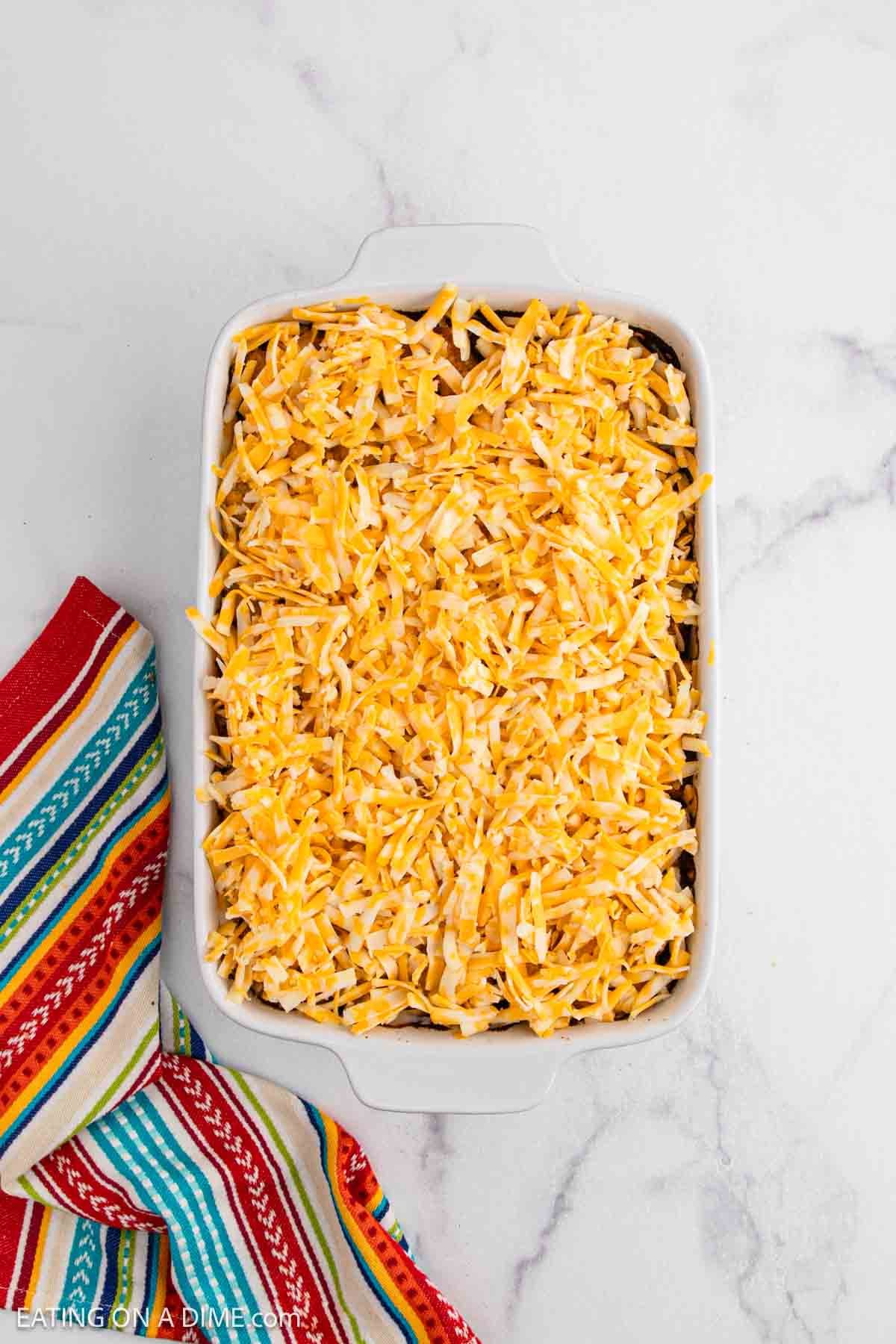 A white casserole dish filled with a layer of shredded yellow and white cheese, showcasing a delicious Mexican Tator Tot Casserole, sits on a marble surface next to a colorful striped cloth.