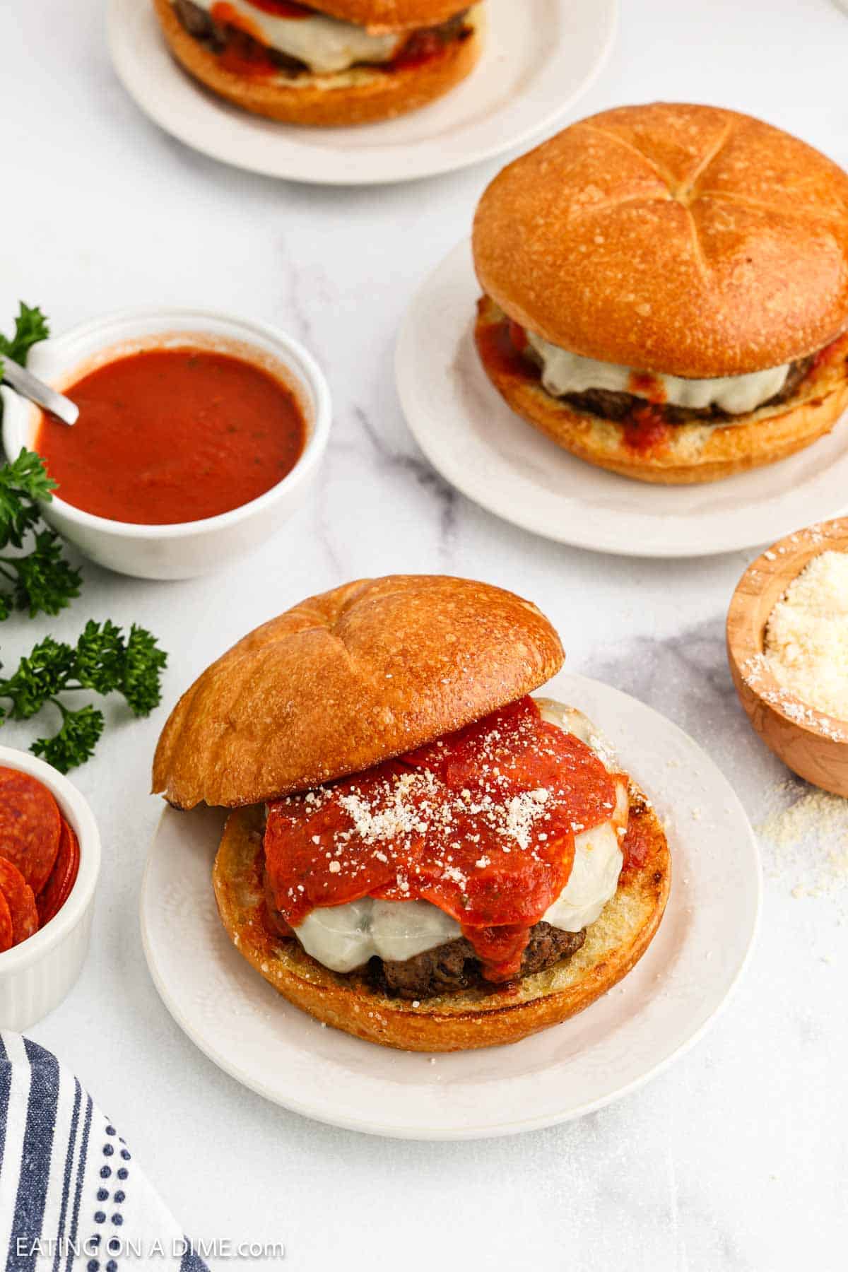 A plate with a sandwich featuring melted cheese and marinara sauce on a toasted bun, surrounded by bowls of marinara sauce, grated cheese, and pepperoni slices for a pizza-inspired burger experience on a white surface.