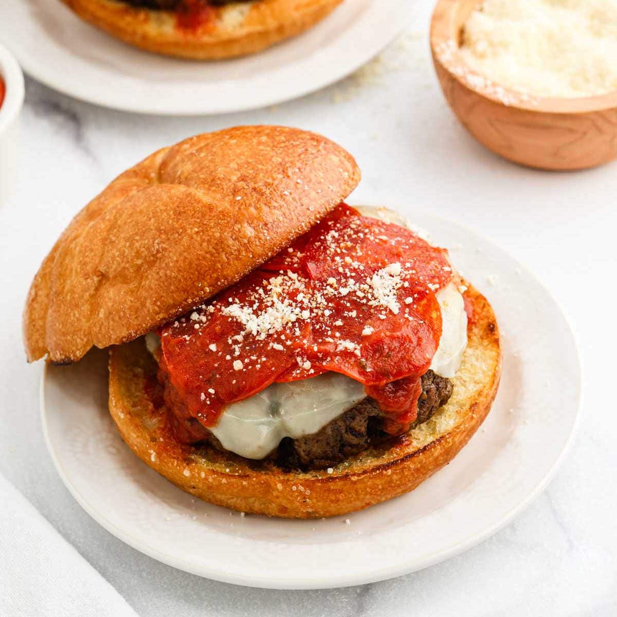 A Pizza Burger on a white plate features a beef patty topped with melted cheese, marinara sauce, and a sprinkle of grated Parmesan on a toasted bun. A small bowl of grated cheese sits in the background.