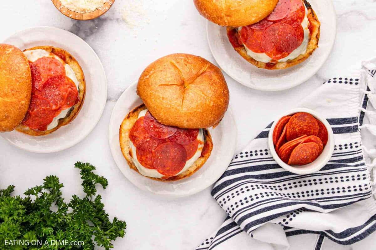 Three plates with delicious Pizza Burgers on sandwich buns sit on a white marble surface. Nearby, a bowl of pepperoni slices and fresh parsley add flavor, while a black and white striped kitchen towel completes the scene.