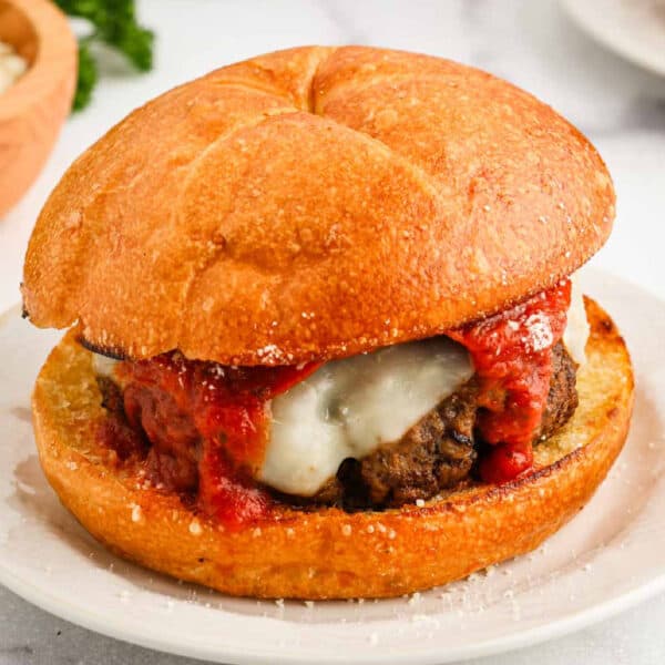 A Pizza Burger featuring a golden, toasted bun, a beef patty topped with melted cheese and zesty marinara sauce, served on a white plate.