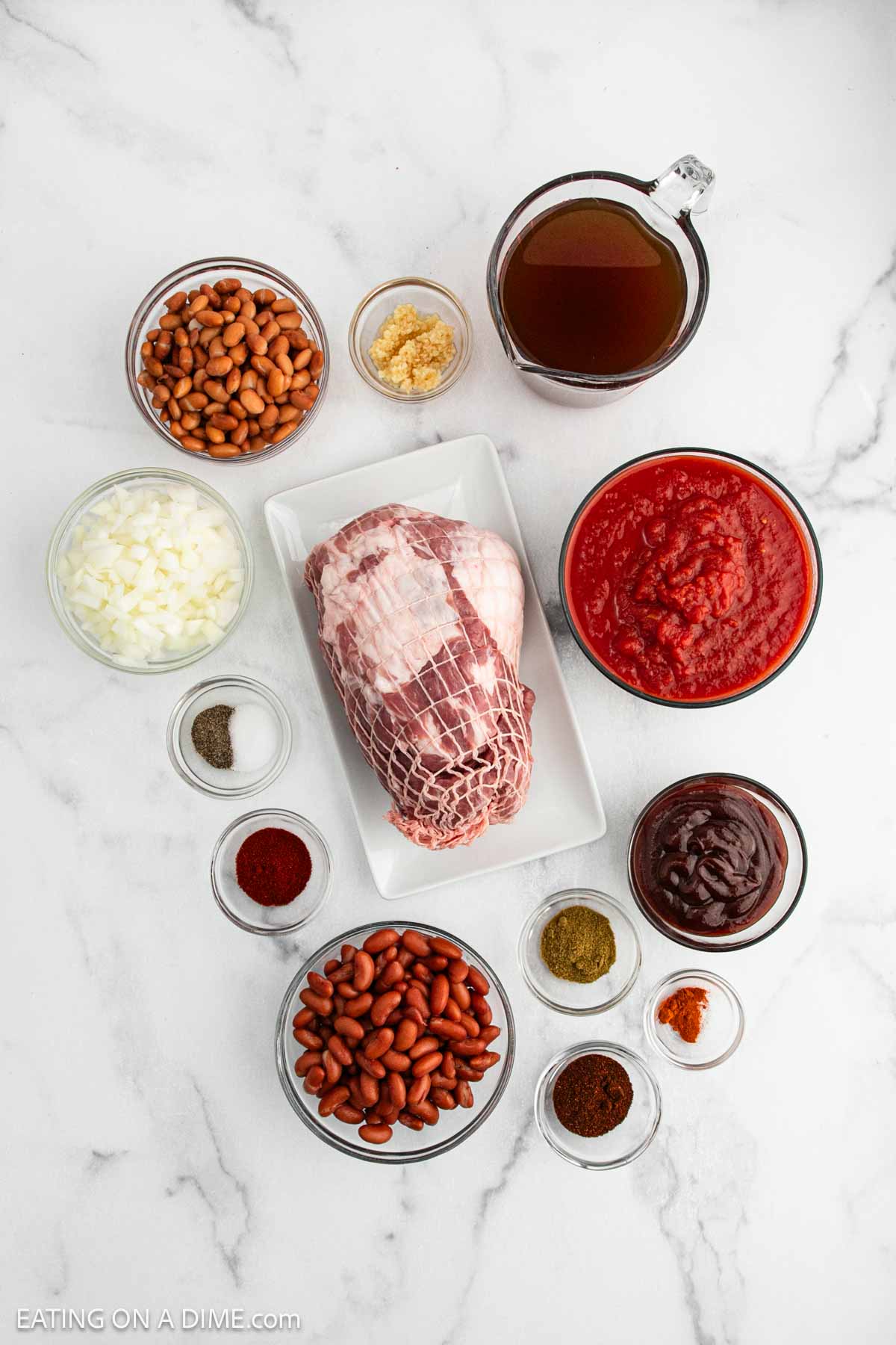 Top-down view of ingredients for Pulled Pork Chili on a white surface, featuring a netted raw pork roast, kidney and pinto beans, chopped onion, minced garlic, tomato sauce, broth, and an array of spices in small bowls.