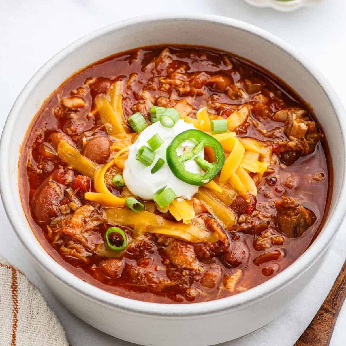 A bowl of Pulled Pork Chili topped with shredded cheese, sour cream, sliced jalapeños, and chopped green onions sits on a white surface. The chili looks thick and hearty, packed with visible beans and juicy chunks of pulled pork.