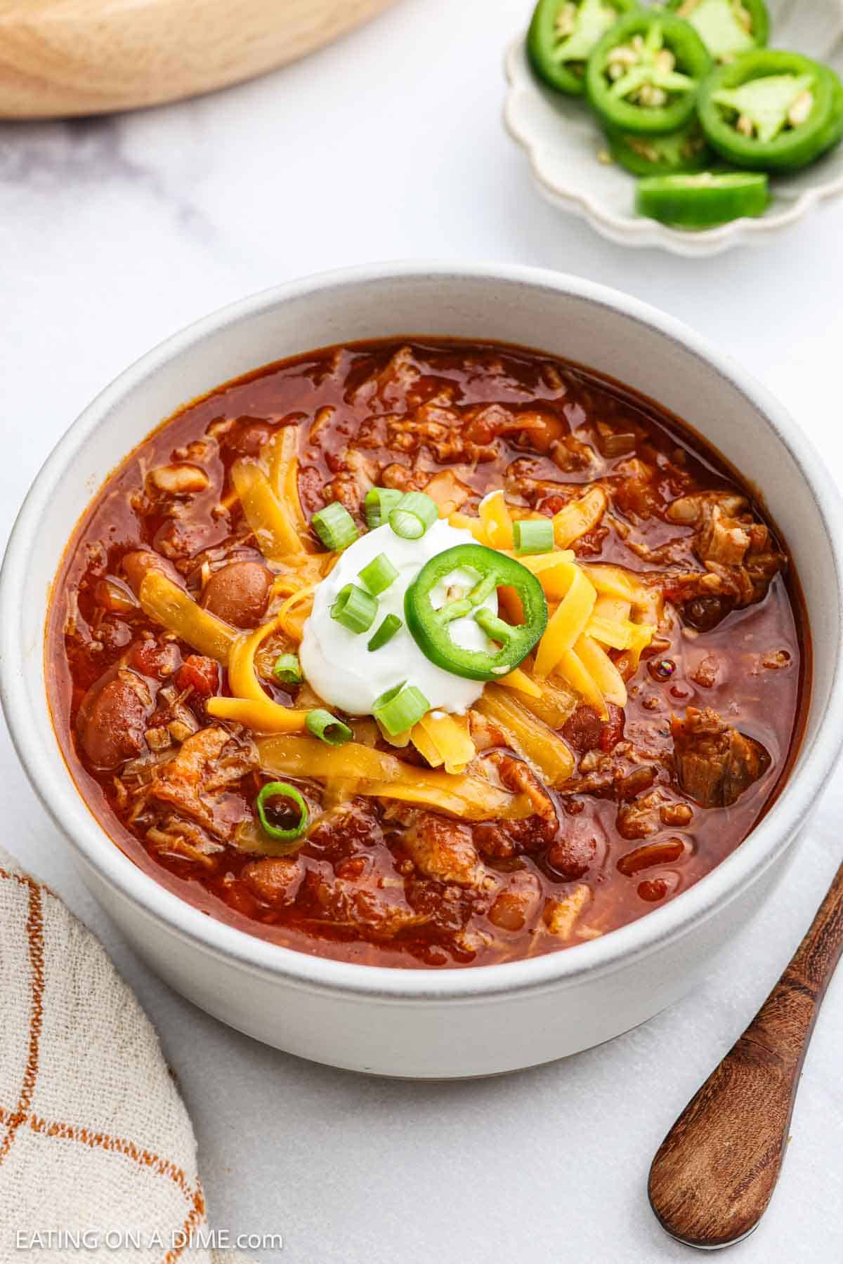 A bowl of Pulled Pork Chili topped with shredded cheese, sour cream, sliced green onions, and jalapeño slices, with a small plate of jalapeños and a napkin nearby.