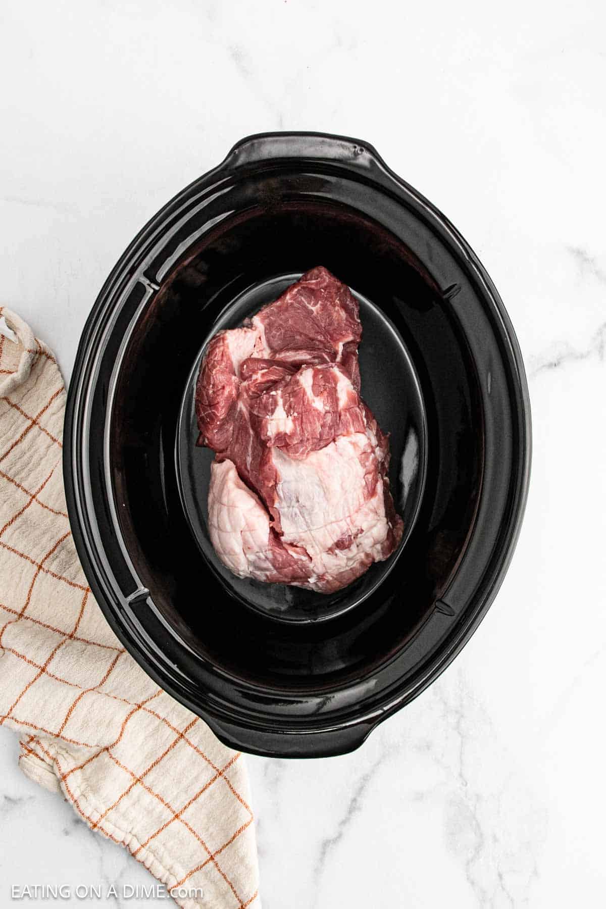 Raw pork shoulder placed inside a black slow cooker, perfect for making tender pulled pork, with a beige and orange checkered kitchen towel nearby on a white marble surface.