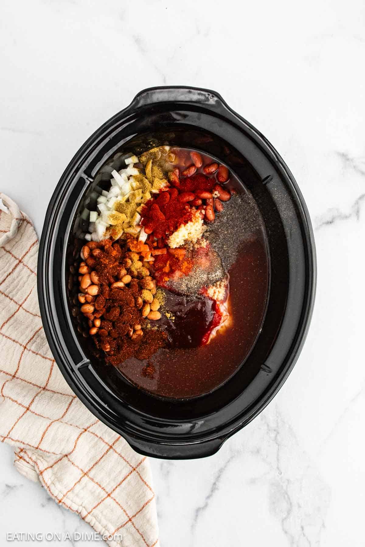 A slow cooker filled with raw Pulled Pork Chili ingredients, including beans, chopped onions, spices, and tomato sauce, arranged in separate piles on top of raw meat. A beige checkered towel lies beside the slow cooker on a marble surface.
