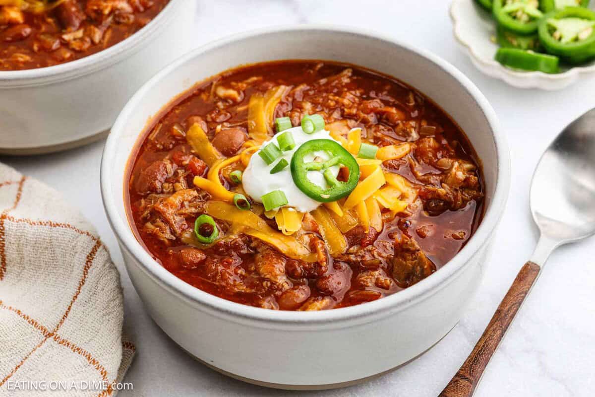 A bowl of pulled pork chili topped with shredded cheddar cheese, sour cream, sliced jalapeños, and chopped green onions. A spoon, a napkin, and a small plate of sliced jalapeños are nearby.