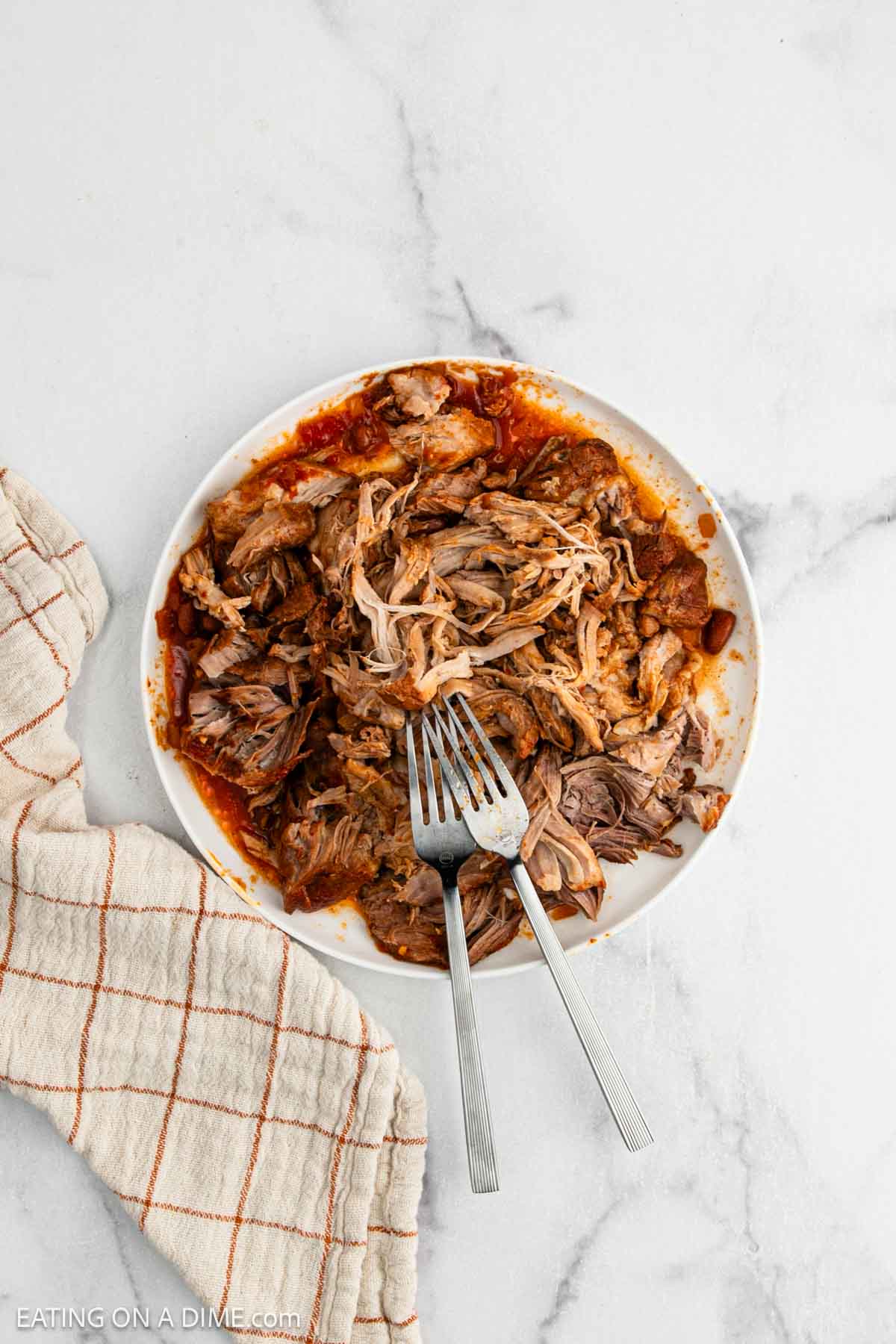A plate of tender shredded Pulled Pork Chili with two forks rests on a white marble surface. A beige and red plaid kitchen towel is placed next to the plate.