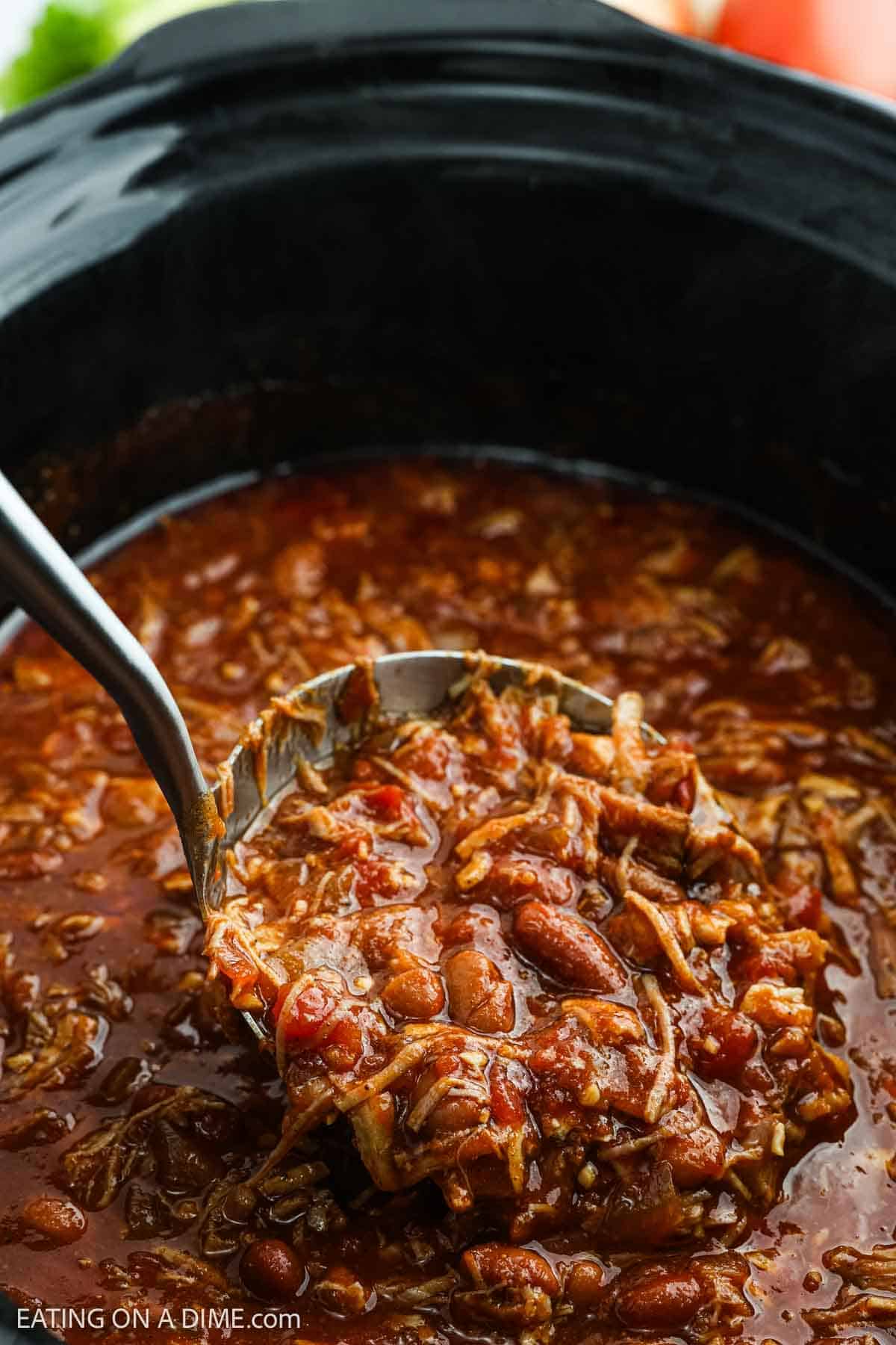 A ladle lifts a serving of hearty Pulled Pork Chili with beans and shredded meat from a black slow cooker filled with thick, rich tomato-based sauce.